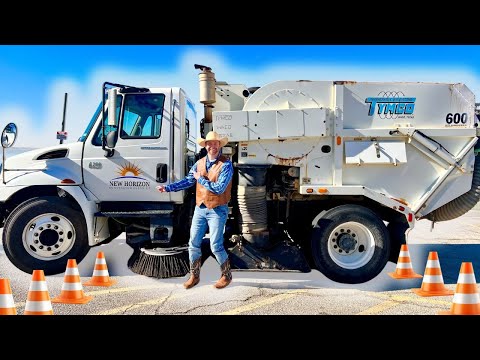 Road crew worker wearing a hat and vest stands beside a white Tymco street-sweeper truck on a sunny road, with orange traffic cones in front.