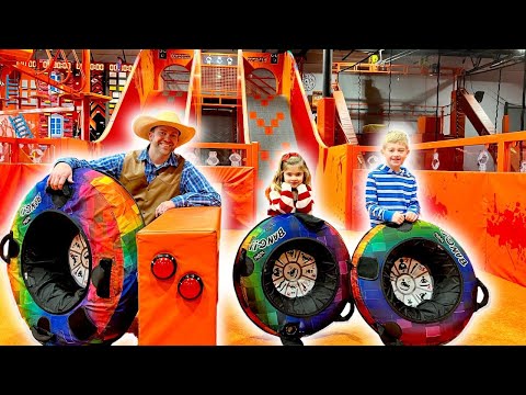 Man in a cowboy hat and blue plaid shirt stands beside rainbow-colored inflatable tires with two children in a bright trampoline park.