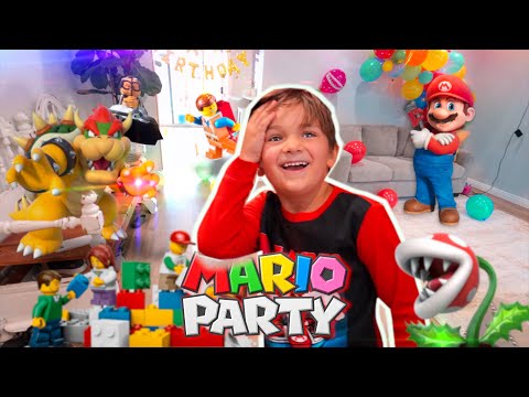 Smiling boy in red and black at a Mario Party birthday party with a large Mario figure, Bowser statue, balloons, and colorful toy bricks.