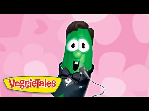 Green VeggieTales cucumber in a tuxedo, bow tie, and microphone, performing against a pink patterned background.