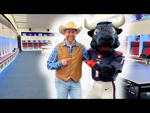 Smiling man in a cowboy hat and brown vest posing with a bull mascot in a bright locker room.