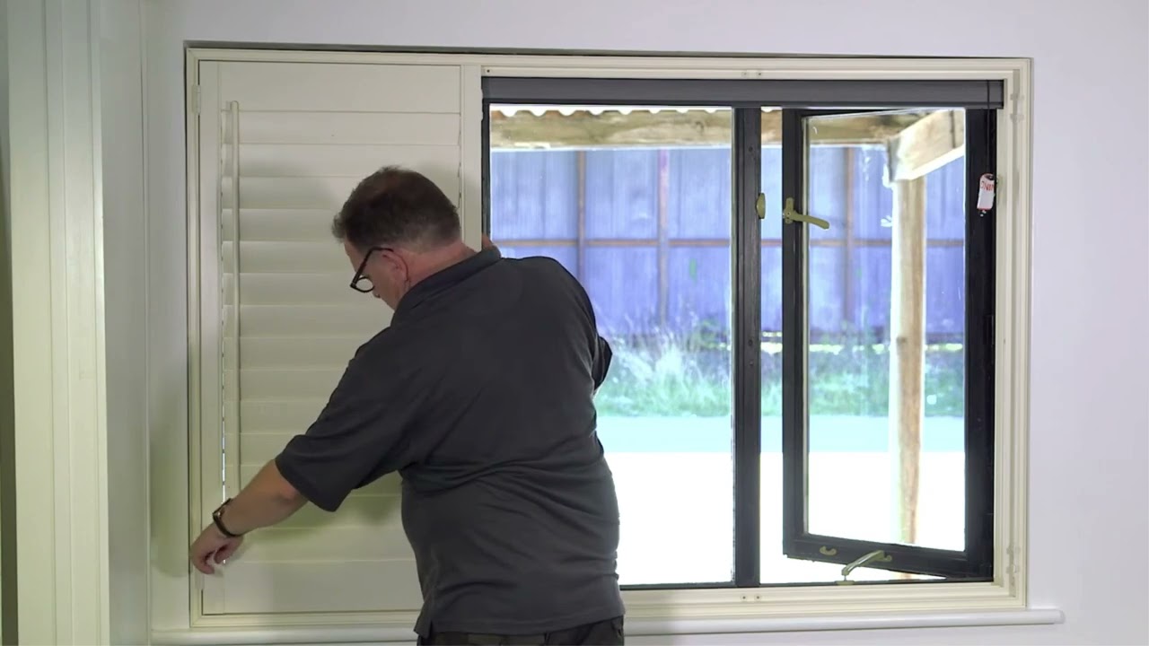 A fitter hanging a white shutter panel in a window frame