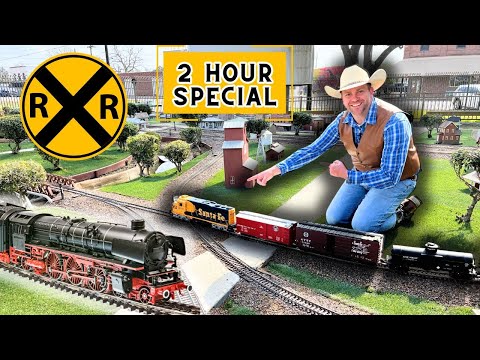Man kneeling beside a detailed model railroad layout with trains, tracks, and miniature buildings, under a bright '2 HOUR SPECIAL' sign and a large yellow RR crossing symbol.