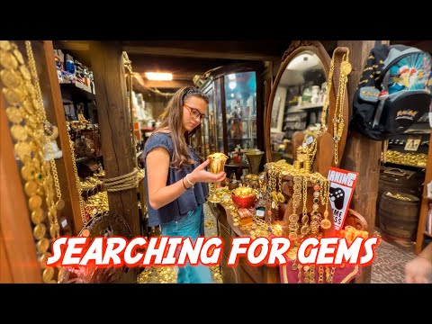 Young woman with glasses holds a small gold goblet in a treasure-filled jewelry shop, surrounded by gold coins, necklaces, and shelves of jewelry.