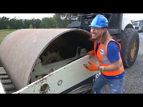 Construction worker in a blue hard hat and orange safety vest smiling beside a large road roller outdoors on a gravel lot.