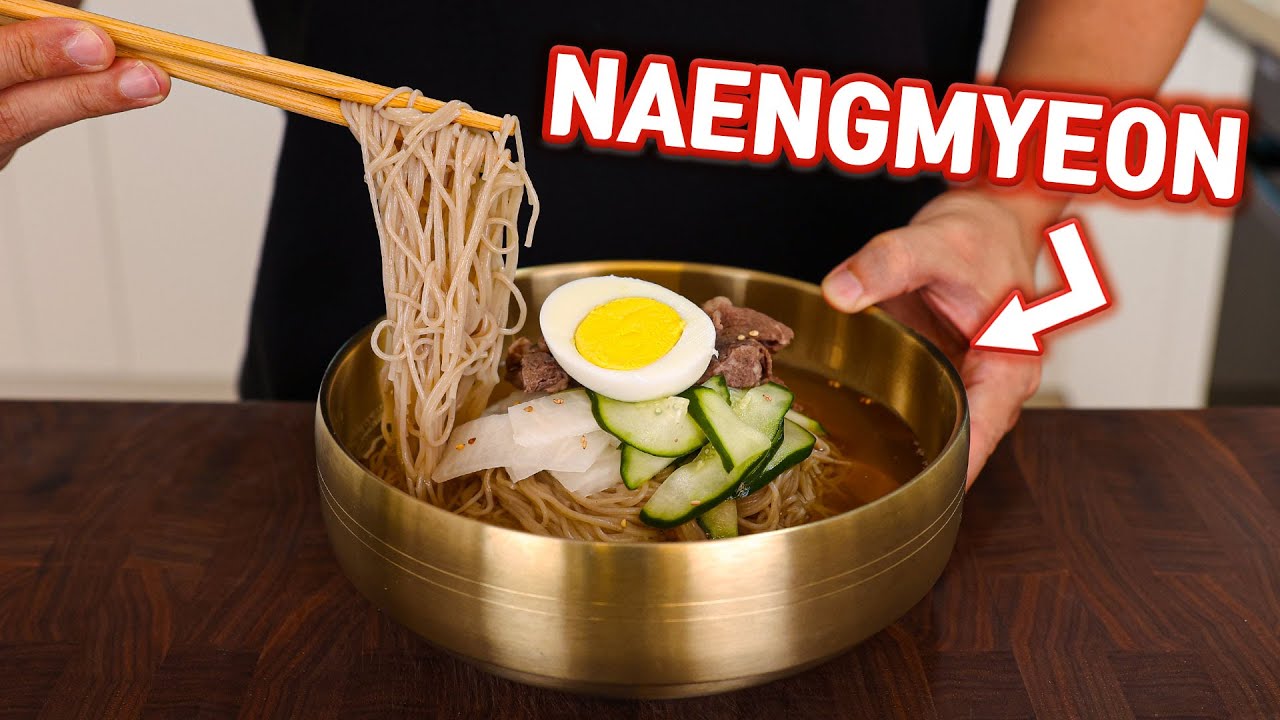 A bowl of Korean mul naengmyeon cold noodle soup with ice cubes clear broth cucumber and beef slices arranged for a summer meal