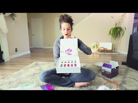 Woman sitting cross-legged on a living room rug opens a white gift box revealing multiple colorful nail polish bottles, surprised expression on her face.
