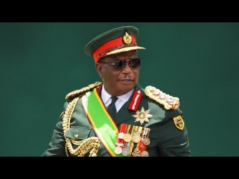 Vice President Constantino Chiwenga speaking at a press conference in Harare, with a stern expression, podium and Zimbabwe flags in background.