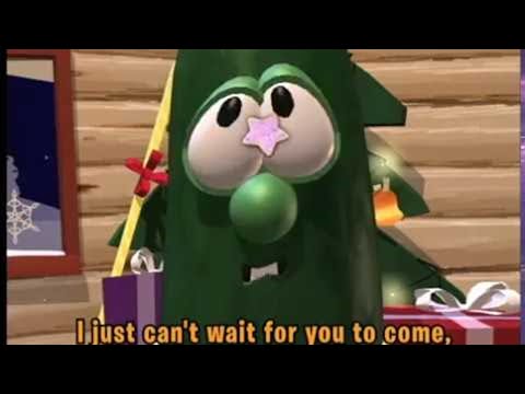 Cartoon green creature dressed as a Christmas tree with presents around, looking toward the viewer.