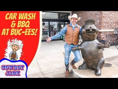Man in a cowboy hat posing with a bronze cowboy statue outside Buc‑ee's beside a red sign that reads 'Car Wash & BBQ at Buc‑ee's' and 'Cowboy Jack'.