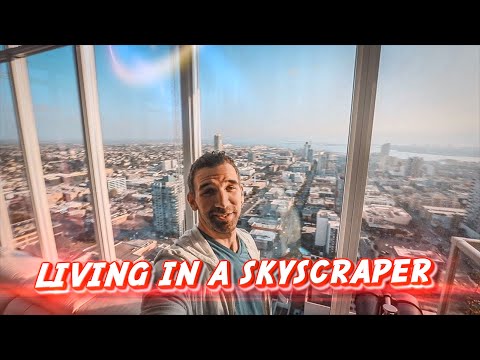 Man selfie in a glass-walled high-rise with a city skyline; red caption 'LIVING IN A SKYSCRAPER' across bottom.