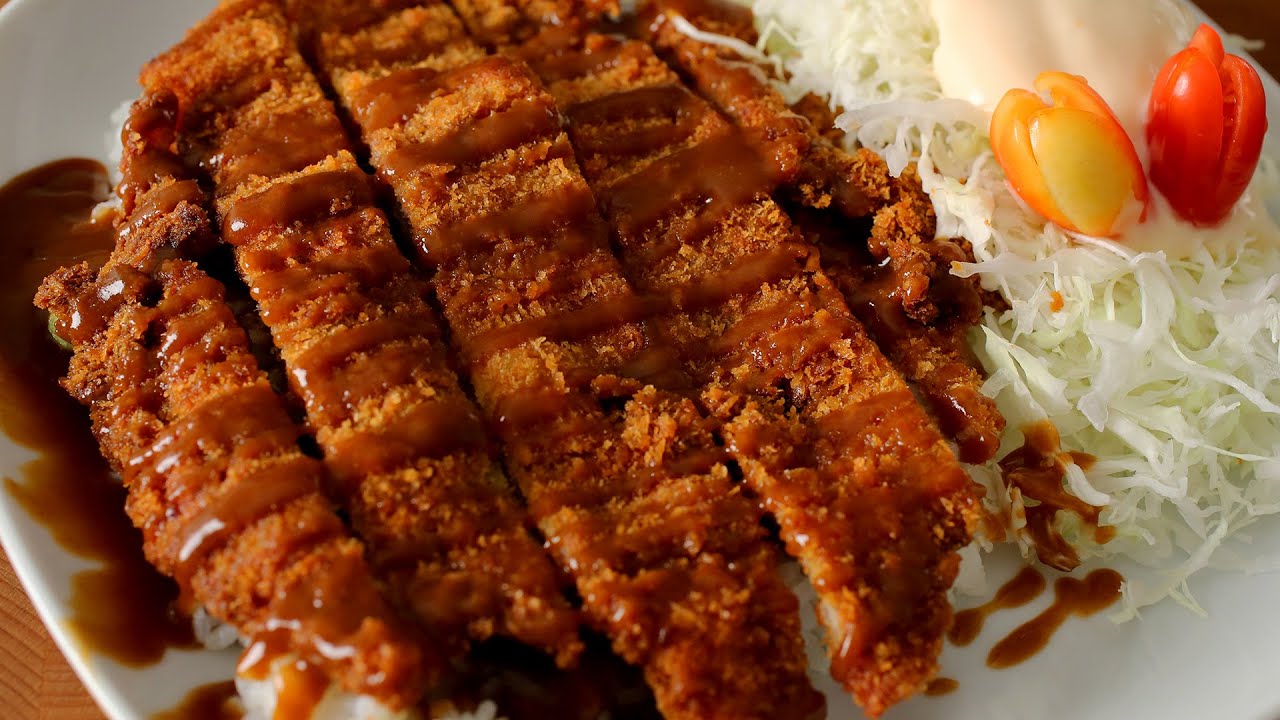 A large Korean donkkaseu pork cutlet covered in crispy golden breadcrumbs served on a plate with rice cabbage salad and brown donkkaseu sauce