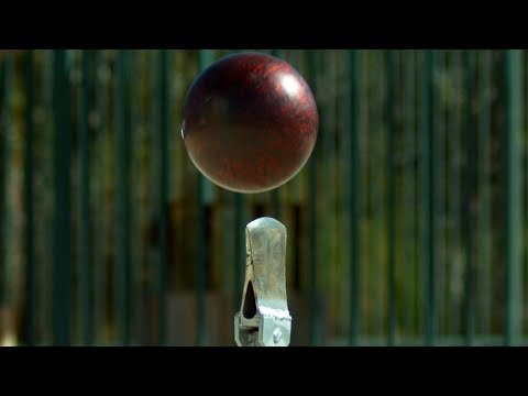 Red cricket ball suspended in mid-air above a metal stump, with a green fence background, conveying a wicket action moment.