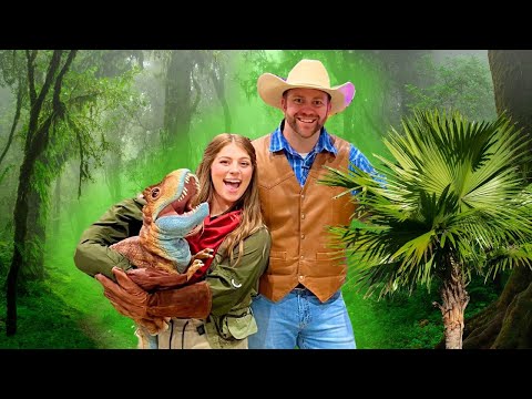 Woman in a green jacket holding a colorful dinosaur puppet, smiling with a man in a brown vest and cowboy hat in a jungle backdrop.