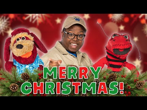 Person in safari-style outfit with two puppets, 'Merry Christmas' text in front, festive red background with pine garland and pinecones.