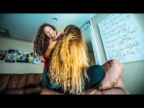 Woman with curly hair cuts another person’s long blonde hair in a casual living room scene, sitting on a brown couch by family photos and artwork.