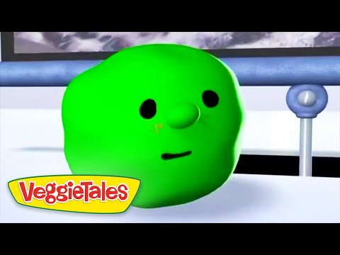 Close-up of a bright green cucumber character with black eyes and a small mouth, from VeggieTales, resting on a white surface with a logo in the corner.