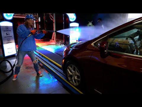 Worker wearing a blue jacket and helmet uses a high-pressure nozzle to spray a car at a car wash bay, colorful lights around.