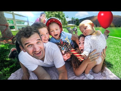Family selfie on a sunny day at the park with kids, a baby wearing a helmet, and a red balloon nearby.