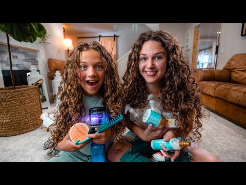 Two girls with long curly hair sit on the living room floor, smiling, holding assorted hair care products and brushes.