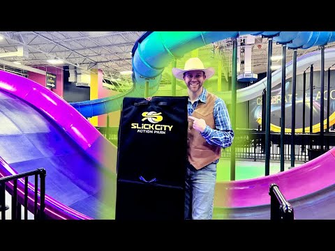 Man in a cowboy hat and vest holds a black Slick City Action Park bag beside colorful indoor slides in the background.