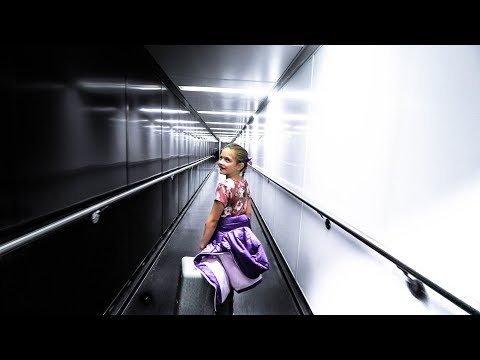 Young girl in a floral top with a purple jacket and small suitcase walks through a bright airport jet bridge, looking back over her shoulder.