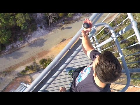 Person standing on a tall metal railing on a tower, holding a rock toward the edge with a ground path visible below.
