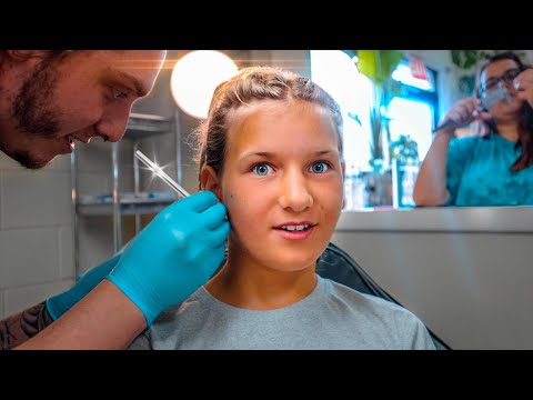 Girl in a dental chair receives a local anesthesia shot from a dentist wearing blue gloves; a friend or assistant is in the background.
