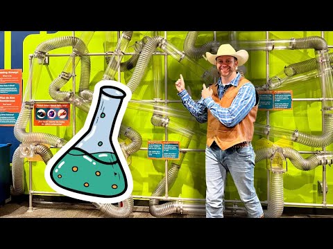 Smiling man in a cowboy hat and brown vest points at a large cartoon flask in a bright lab exhibit with tangled metal tubes behind him.