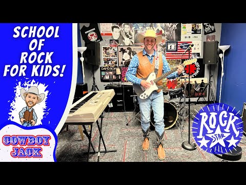 A man wearing a cowboy hat and vest plays a white electric guitar in a kids’ rock studio, with a large blue ‘School of Rock for Kids!’ banner and posters on the wall behind him (Cowboy Jack).