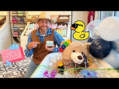 Man in a cowboy hat and brown vest smiles while pointing to a bar of soap in a colorful soap shop display, with a teddy bear in a tub adorned with flowers and balloons nearby.