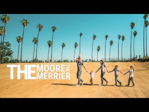Family of six walking hand-in-hand across a sandy hill under tall palm trees, with large 'The Moore Merrier' text on the left (banner photo).