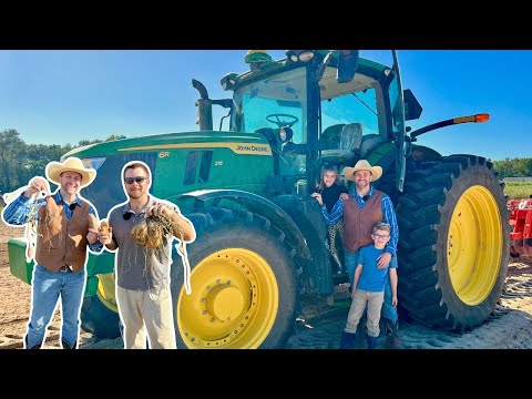 A group of people posing with a large green John Deere tractor with yellow wheels on a sunny farm field; harvest items in their hands suggest a harvest moment.