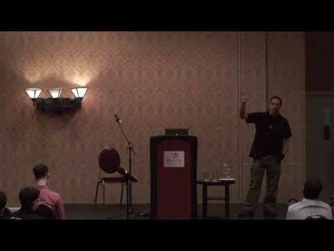 A man in a darkened conference room gives a talk standing to the side of a lectern. The wallpaper is brown-ish patterned hotel style (not expensive) the man's clothes are casual - slacks, short sleeve grey shirt. The video quality is not high definition, looks old (it's from 2012)