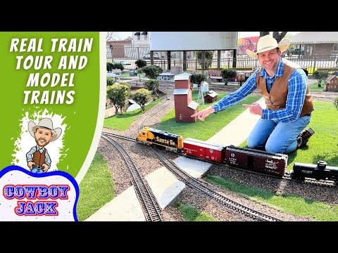 Miniature train layout with a smiling man in a cowboy hat kneeling beside tracks, pointing at the trains; left side shows bold green banner reading 'REAL TRAIN TOUR AND MODEL TRAINS' and a cartoon cowboy with 'COWBOY JACK' logo.