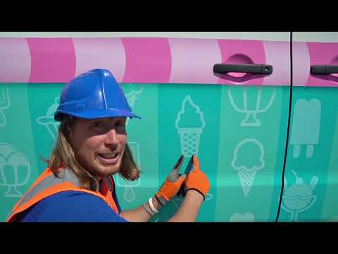 Person wearing a blue hard hat and orange safety vest applying an ice-cream themed vinyl wrap to a teal van with a pink stripe.