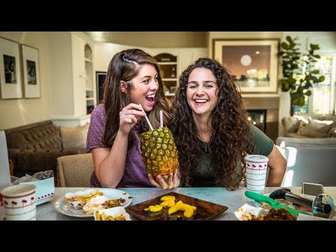 Two women sit at a dining table laughing as one sips from a pineapple with straws held between them in a bright kitchen/living room.