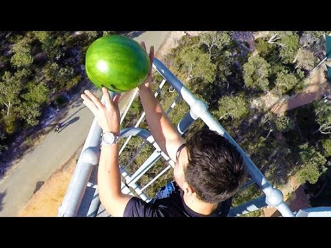 Person on a metal railing high above ground reaching with both hands to place a large green watermelon on the railing.