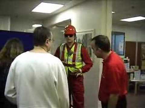 Safety harness competition joke with living mannequin #7 (flash) mannequin-man performming as a Living Mannequin: Fall arrest harness being put onto mannequin man by member of the public at a Safety clothing and PPE exhibition by ARCO at family open day at BAE (British Aerospace Engineering) systems in Rochester #7 (flash) for Arco on 06/07/2002