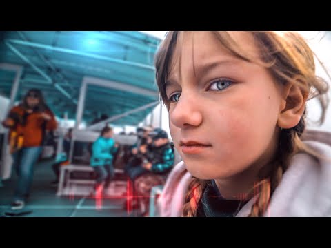 Close-up of a young girl with braided hair and blue eyes, looking at the camera, with a blurred cafe scene in the background under a teal canopy.