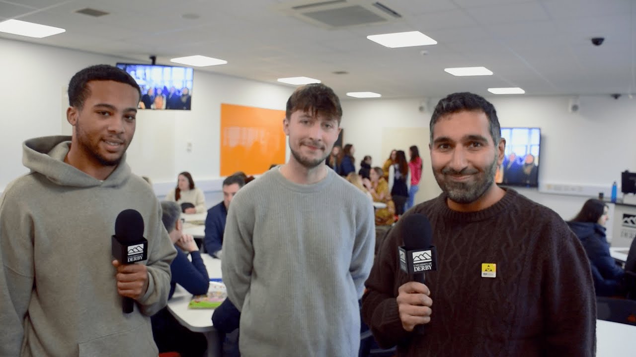 A group of three males in hoodies and casual attire stand with microphones with a busy classroom environment behind them.