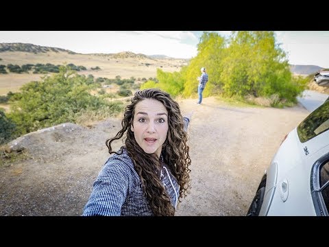 Woman takes a selfie on a dirt road with a white SUV on the right and a man in the distance amid a rural landscape.