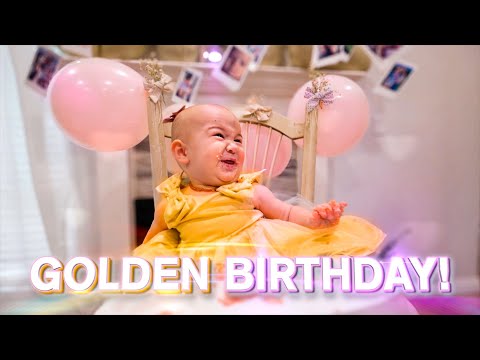 Baby in a yellow dress sits in a small chair at a birthday party with pink balloons in the background and 'GOLDEN BIRTHDAY!' text at the bottom