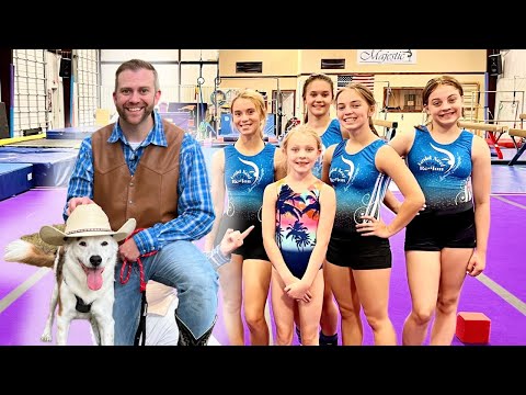Man in a brown vest and blue plaid shirt sits with a white dog wearing a cowboy hat, posing with six young gymnasts in blue leotards in a gym with purple mats.