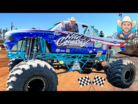 A blue monster truck with oversized tires and a smiling driver in a cowboy hat waves from the window on a dirt track, race flags at the front.