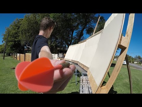 Person extending their arm toward the camera, holding an orange plastic dart while standing beside a curved wooden frame with white panels outdoors on a sunny day.