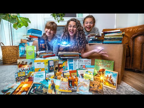 Three people (woman and two children) sit among a colorful pile of children's books on the living room floor, smiling at the camera.