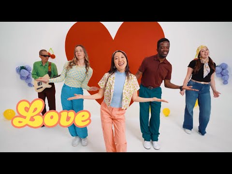 Group of five friends dancing in a bright studio with a large red heart backdrop and retro 'Love' lettering in the foreground; colorful 70s outfits.
