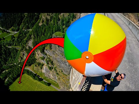 Person in a black shirt holds a giant multicolored beach ball near a cliff edge on a roadway, suggesting a high-risk stunt.