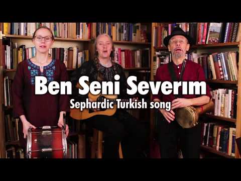 Three musicians standing in front of two bookcases, playing Bulgarian tupan, guitar, and darabuka on “Ben Seni Severım,”, a Sephardic song with lyrics in Turkish and Ladino. This is a still from a video of the Ensemble M’chaiya performing live.